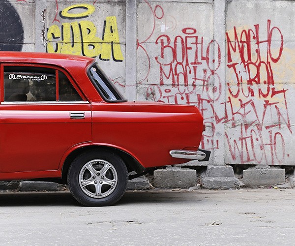 A red car is parked in front of a retaining wall emblazoned with graffiti, including the word Cuba.