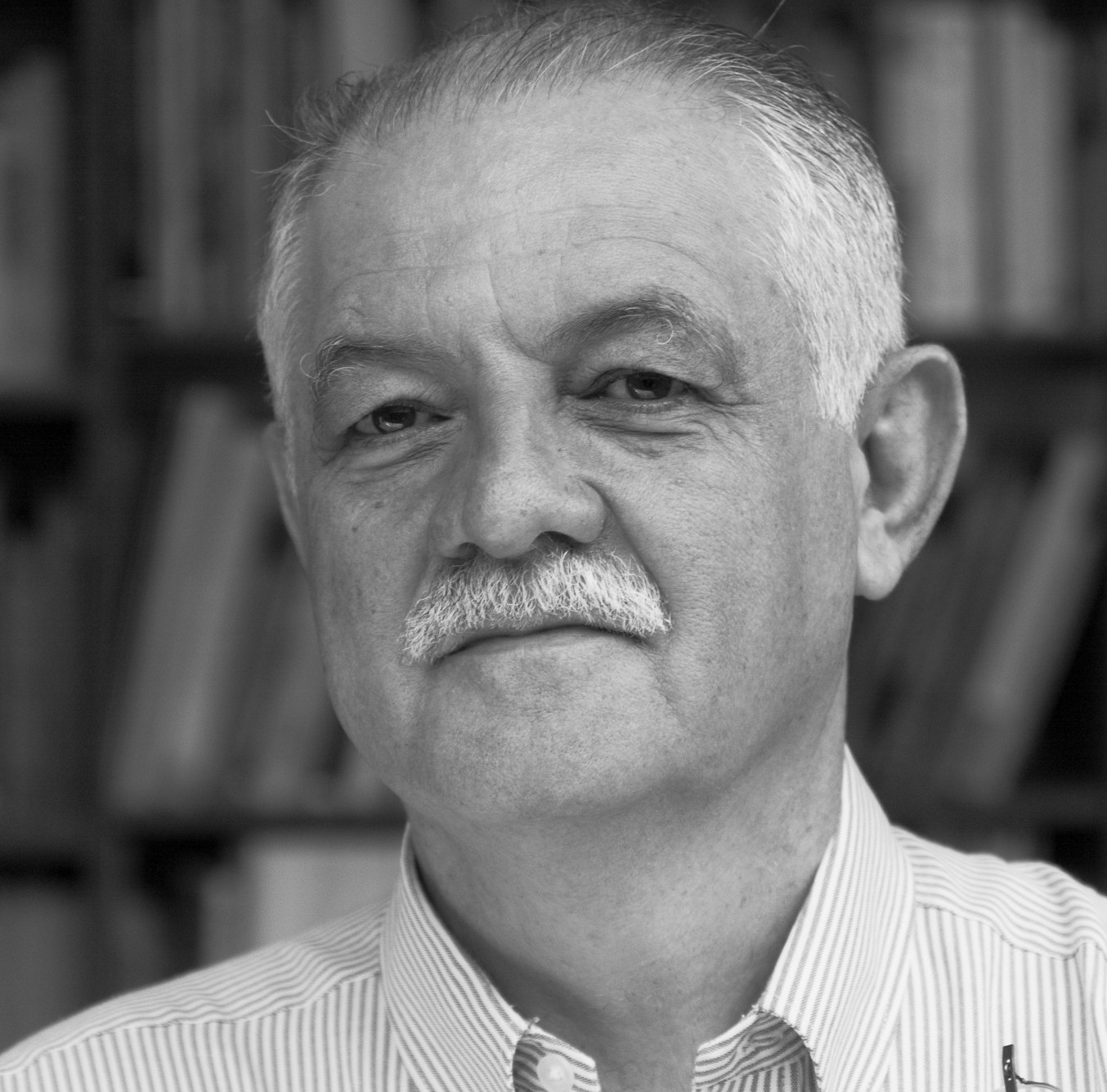 Antonio Azuela is wearing a striped shirt, standing in front of a bookshelf, which is blurred in the picture. The photo is in black and white. 