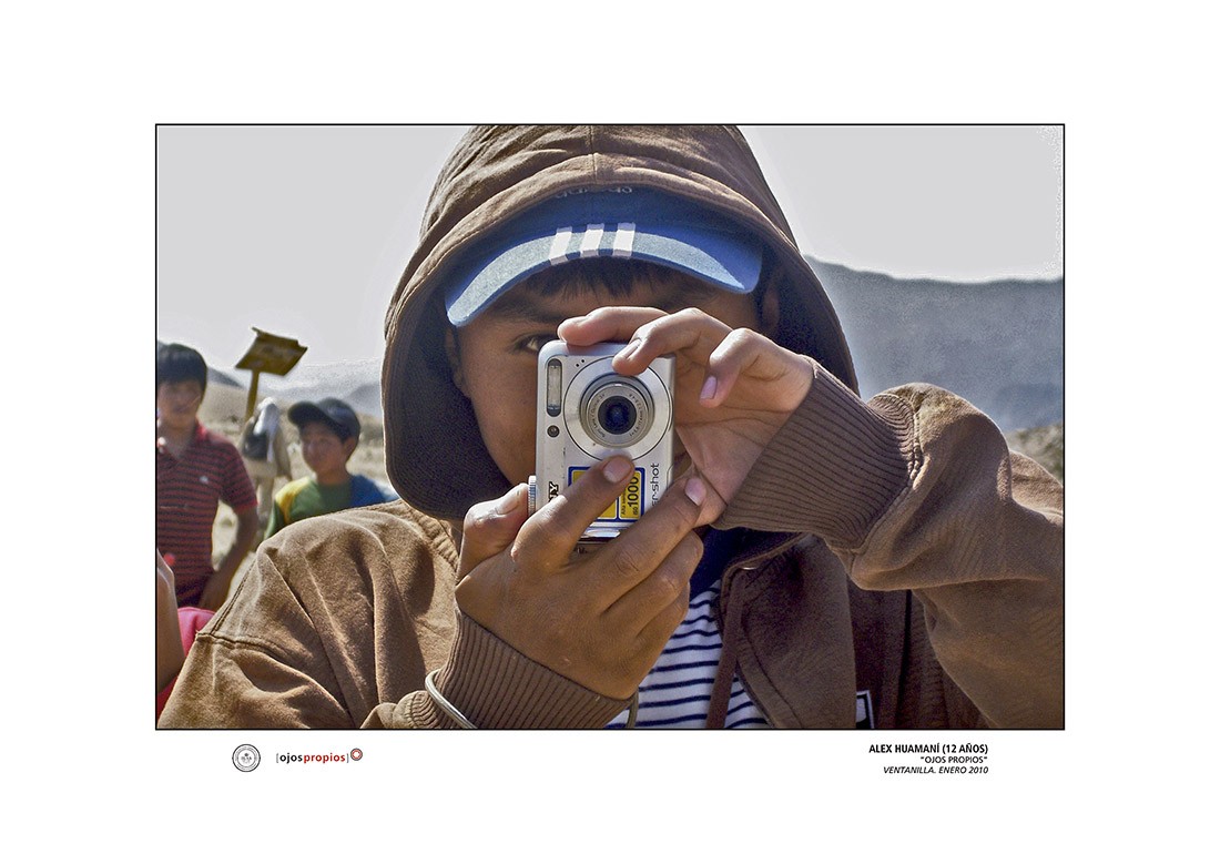 young person wearing a hood and cap holds a digital camera in vertical position while he steadies himself to take a picture