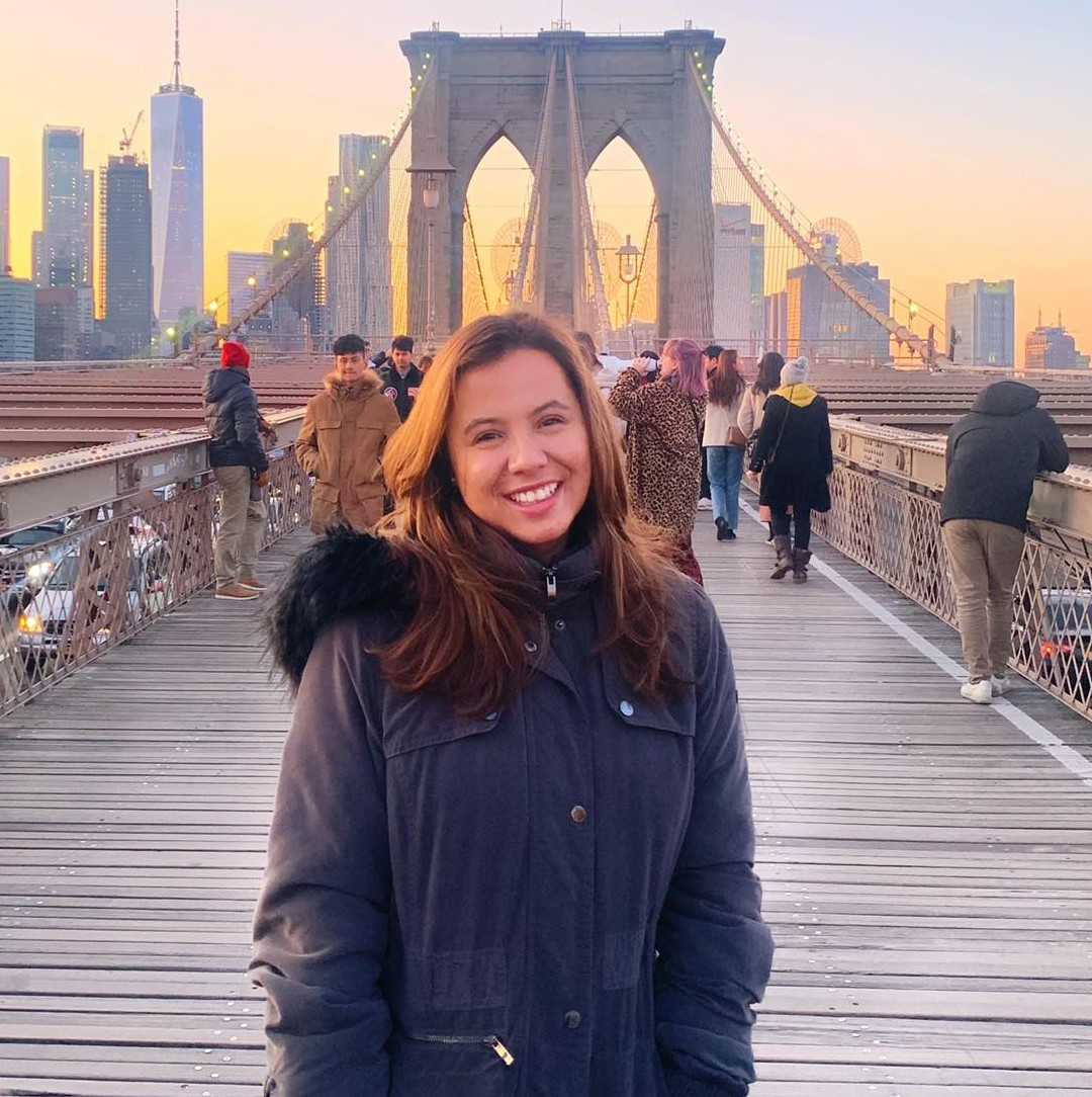 Pilar smiling for the camera in front of a scenic view of a brdige in New York City. She is wearing a black coat.