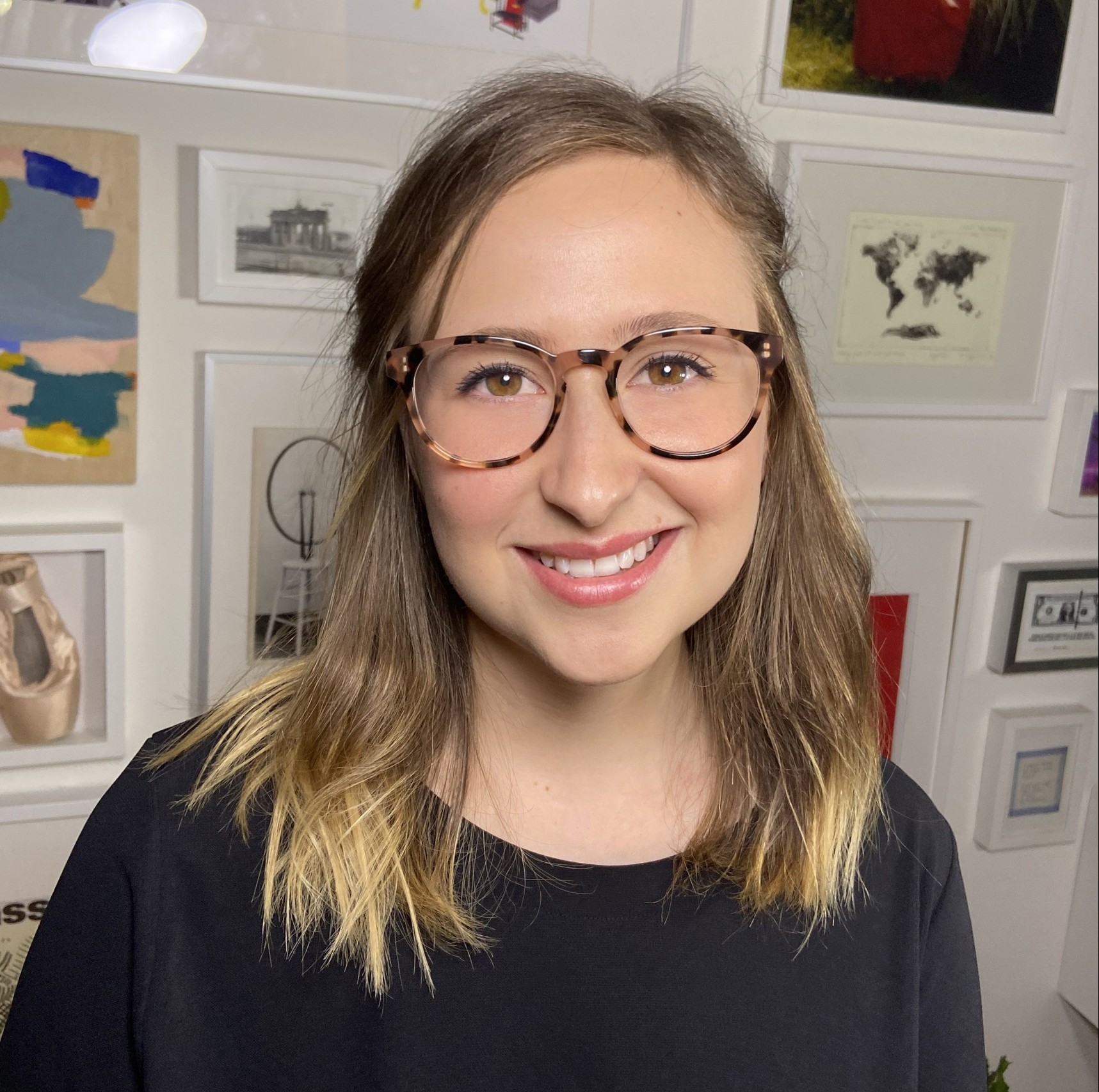 Ana is smiling at the camera in front of a wall with lots of picture frames. She has blond hair and is wearing glasses and a a black shirt. 