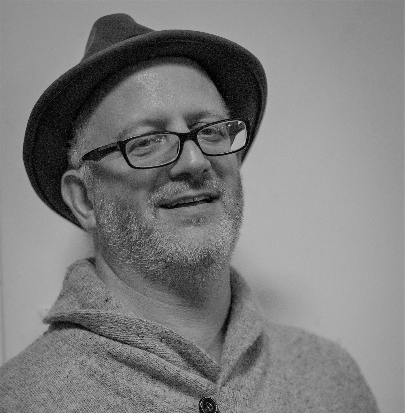 Benjamin is standing against a blank background. He is wearing a hat, a sweater and glasses. The picture is in black and white.