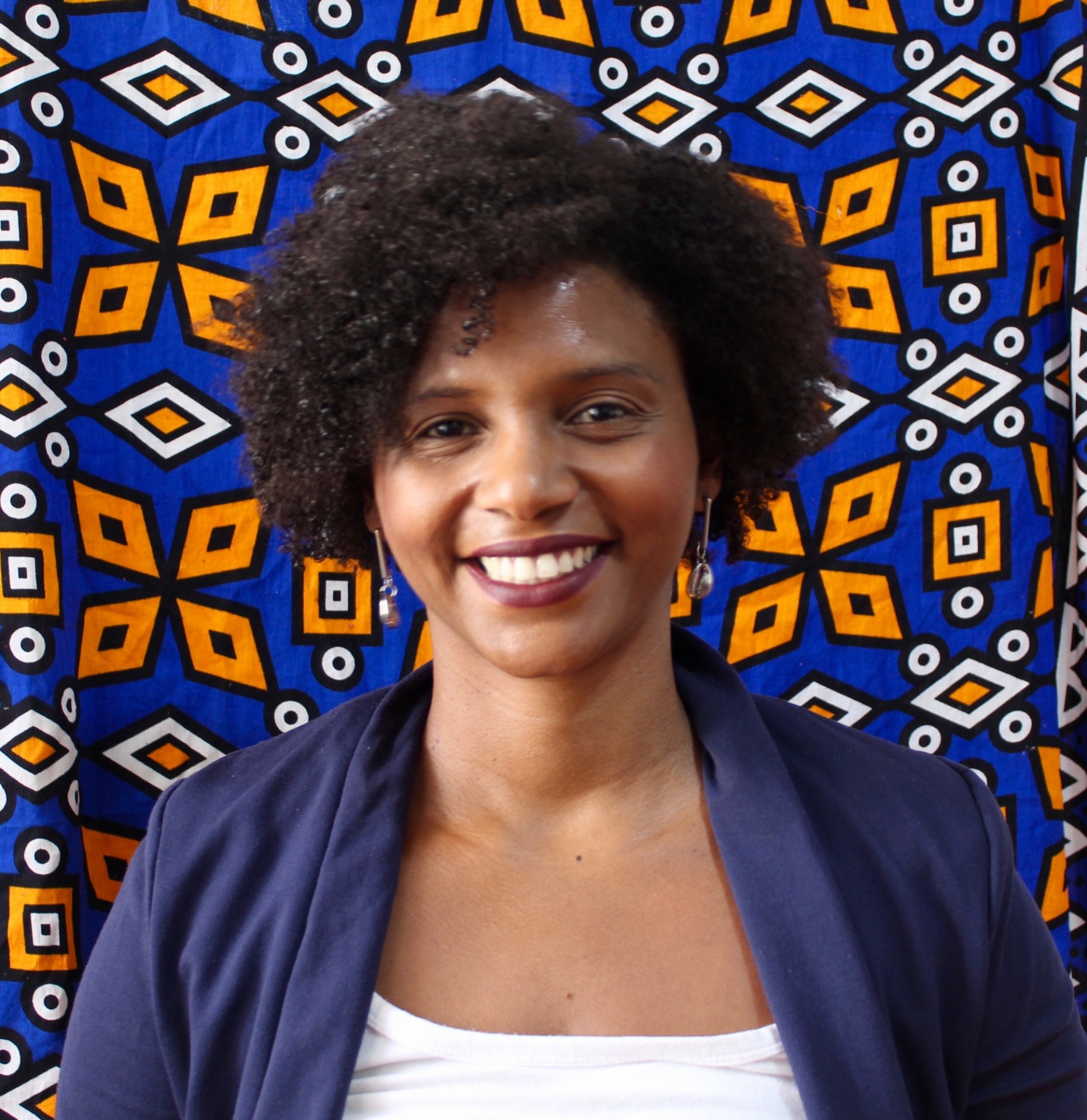 Camila Daniel stands in a white shirt and navy blue jacket against a colorful patterned background.