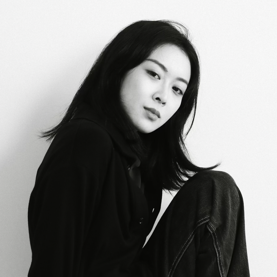Black and white photo of Meiyi standing in front of a white background. She is looking to the side towards the camera; 