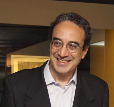 Carlos Augusto Calil is smiling in a suit, unbuttoned light-colored shirt, and glasses.