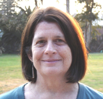 Elsie Rockwell is smiling in a blue t-shirt and silver earrings. In the background there is a patch of grass and trees.