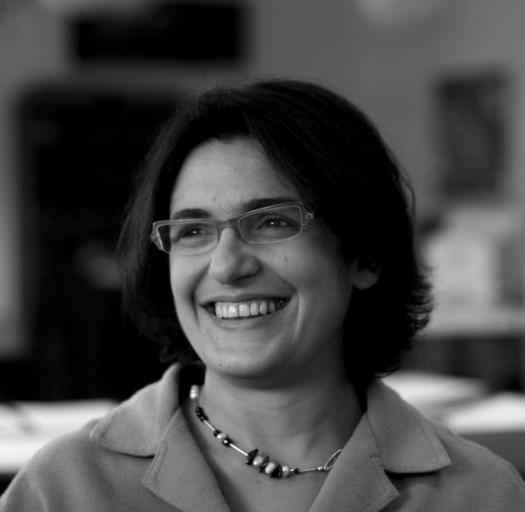 Esther Hamburger is smiling in front of a blurred background. She is wearing a button-down shirt, a collar, and glasses.