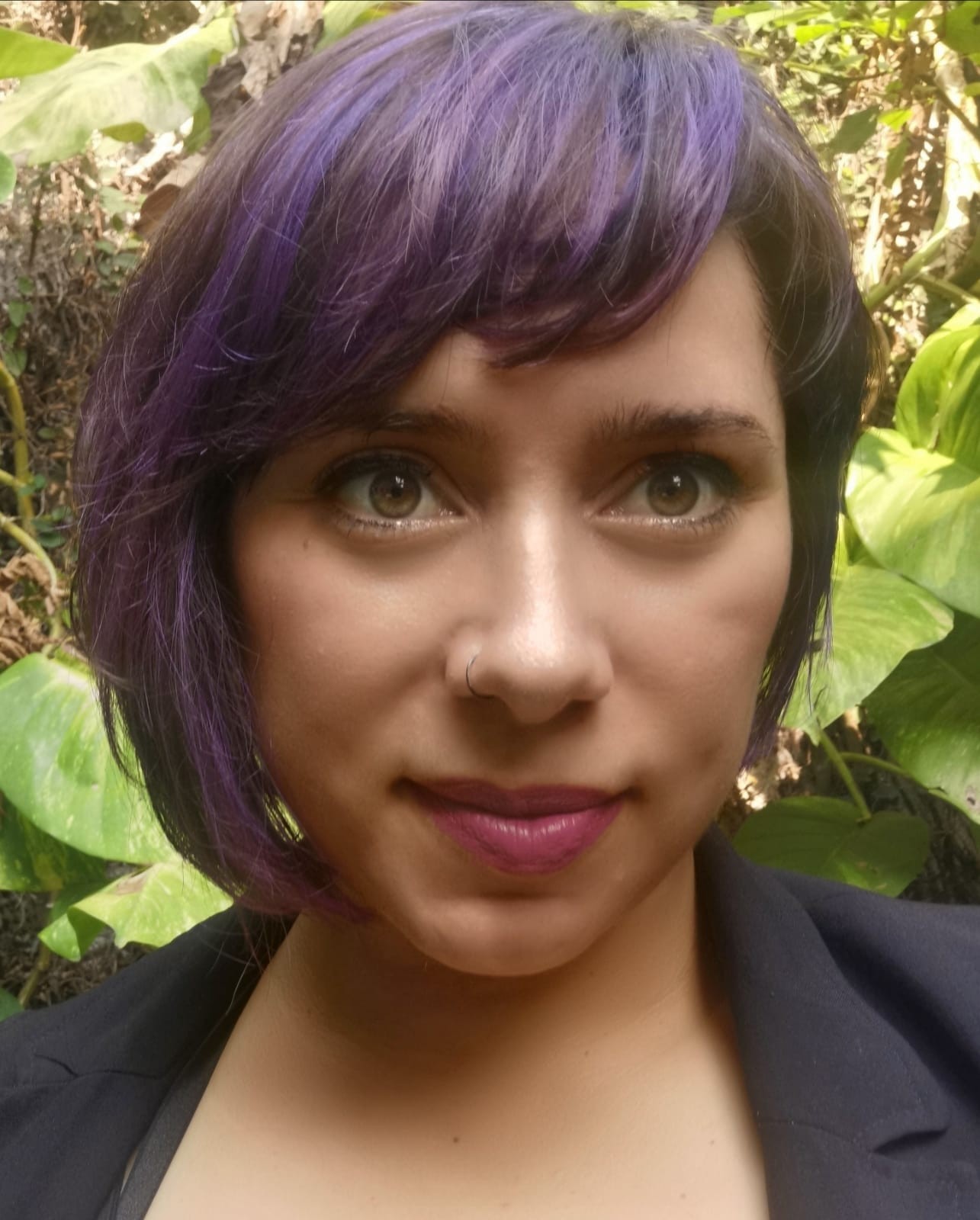 Marisa is standing against a background of wild leaves. She is wearing a black blazer.