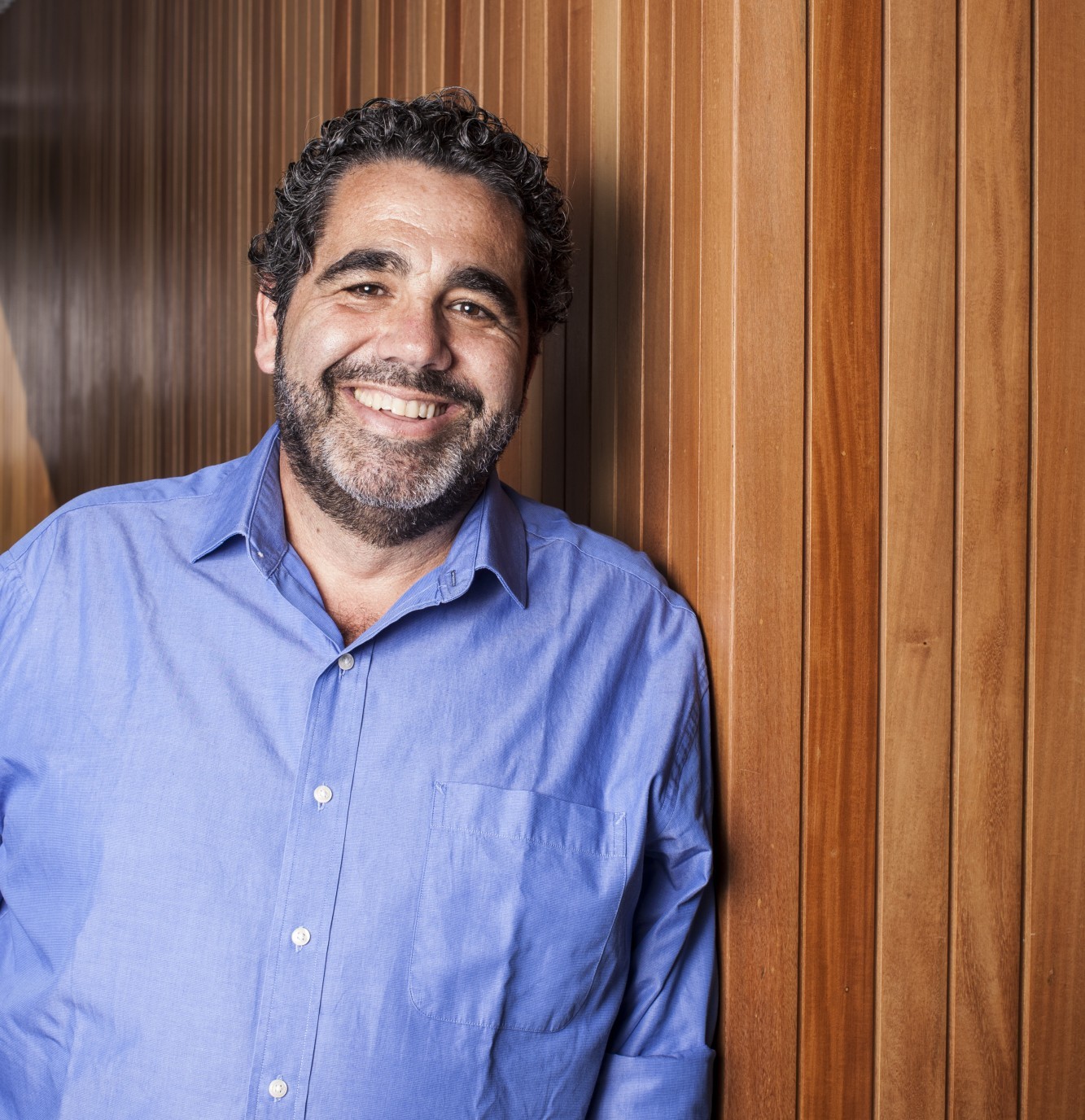 Marcelo is smiling and standing against a background of vertically disposed wood tiles. He is wearing a blue button-down shirt.
