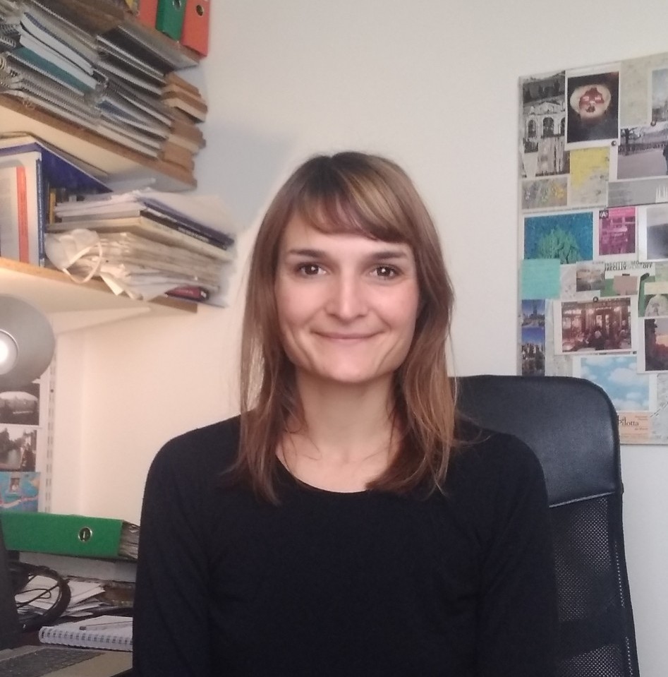 Ines is smiling and sitting in a black chair, wearing a black shirt. In the background, a number of notebooks and folders are on a table and two shelves above it. A number of pictures are posted on a board on the wall to the right.