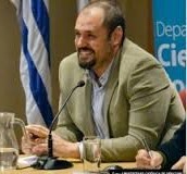 Juan Pablo Luna is smiling while sitting at a conference table. He is wearing a beige blazer and light blue shirt. On the table there is a pitcher of water, a glass filled with water, and a table microphone.