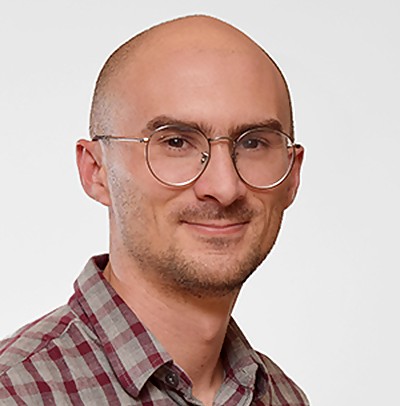 Mauricio is standing against a plain background. He is wearing thin-framed glasses and a plaid maroon-and-gray button-down.