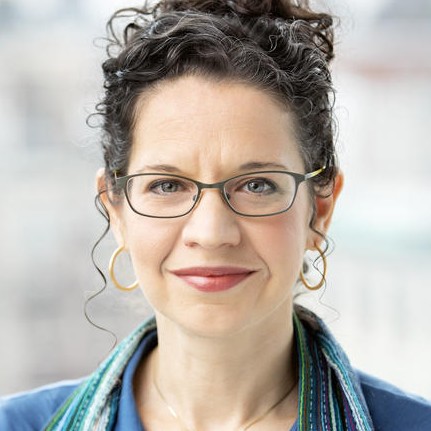 Nara Milanich is wearing a blue shirt and a striped blue and green scarf. She has her curly hair on a bun and is wearing glasses. 