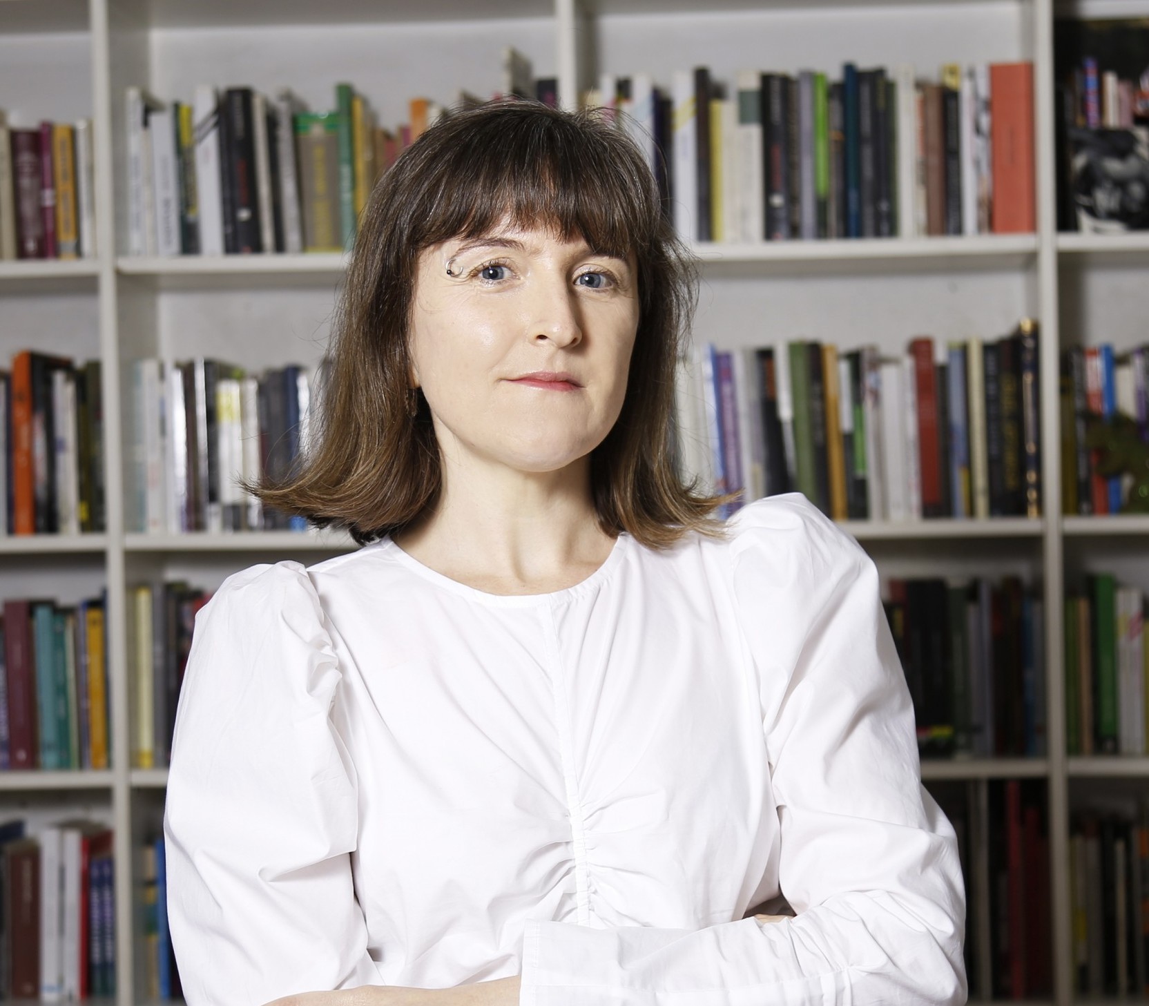 Nayla is standing against a background of books on a white bookshelf. She is wearing a white shirt.