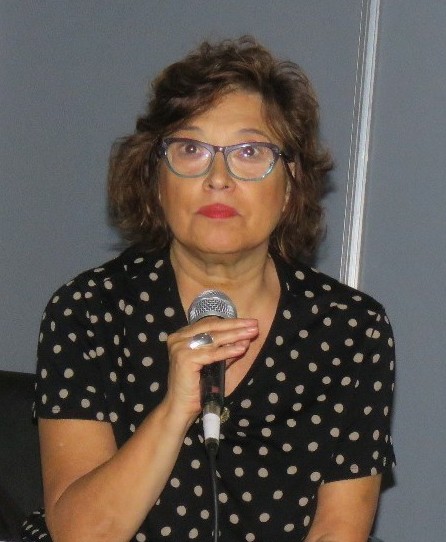 Nora is sitting, holding a microphone while she delivers a message