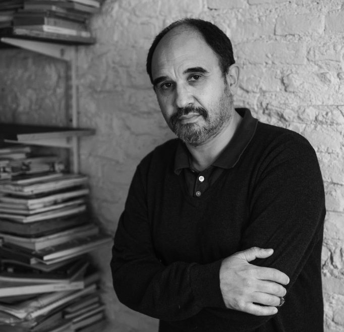 Pablo Pineau stands in jeans and a dark sweater next to bookshelves overflowing with books and journals. The picture is in black and white.