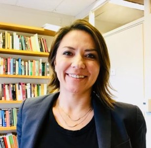 Monica Pachon is smiling in a dark blue blazer, black shirt, and double golden necklace. In the background there are shelves filled with books.