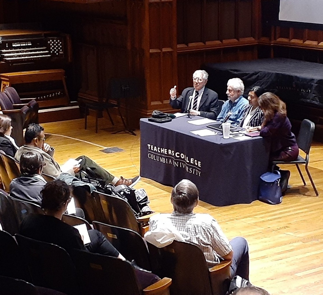 The picture shows a Teachers College conference with four speakers at a table and a full audience in an auditorium.