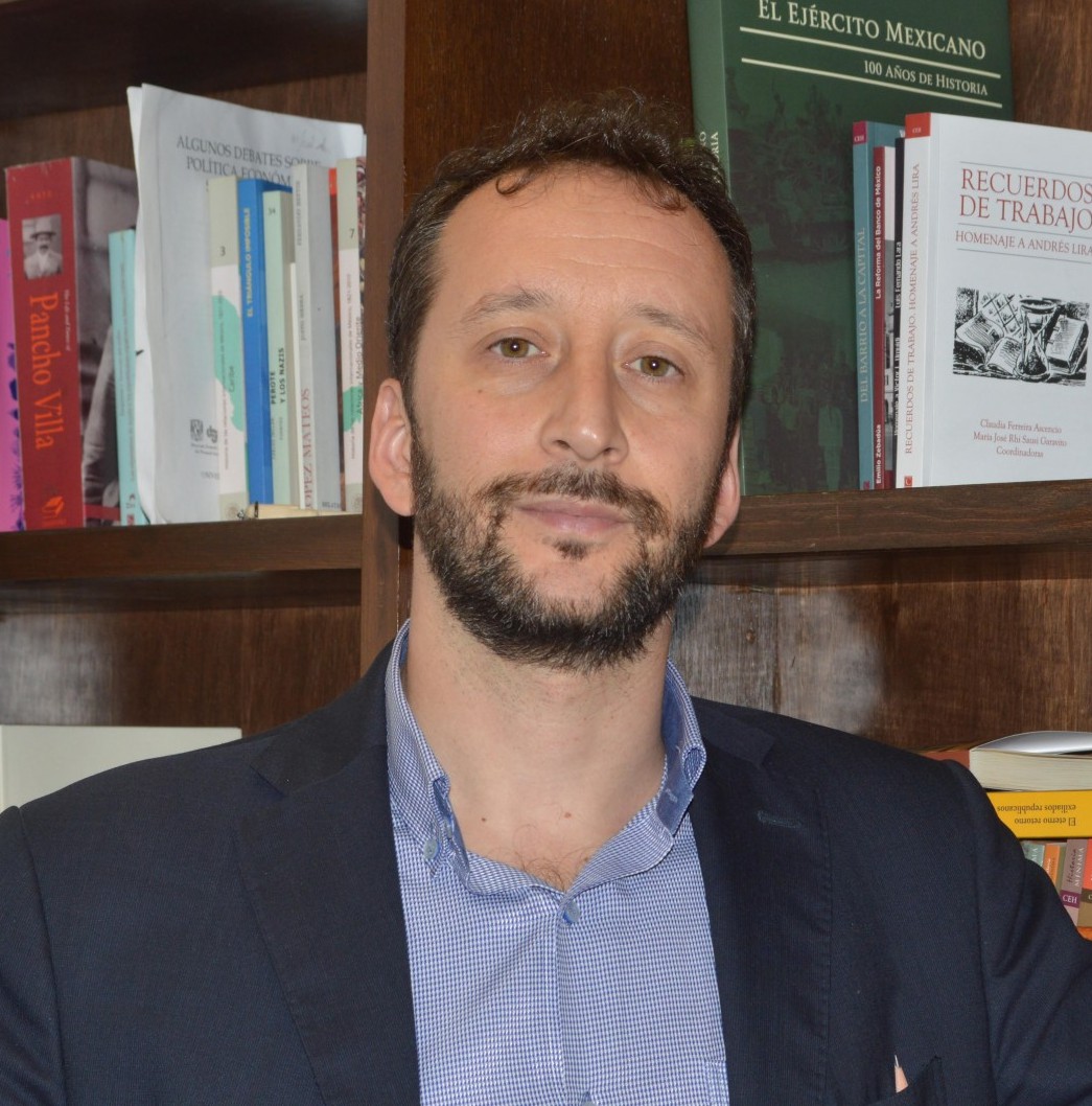 Vanni Pettinà is standing in front of a wooden bookshelf. He is wearing a blue button-down shirt and a dark blazer. 
