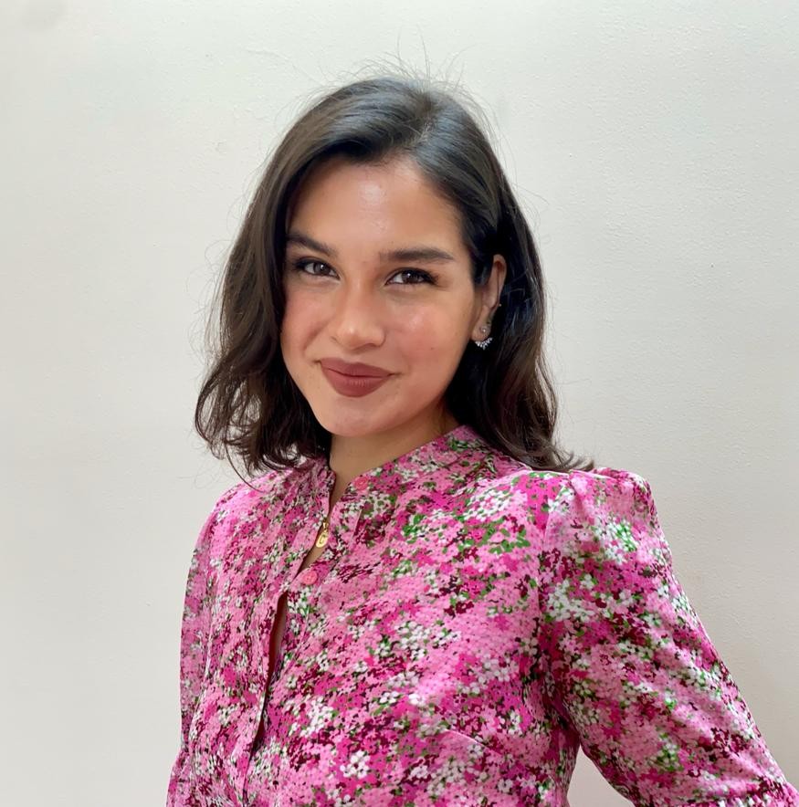 Camila is smiling for the camera as he stands in front of a white wall. She has brown wavy hair and is wearing a pink floral shirt.