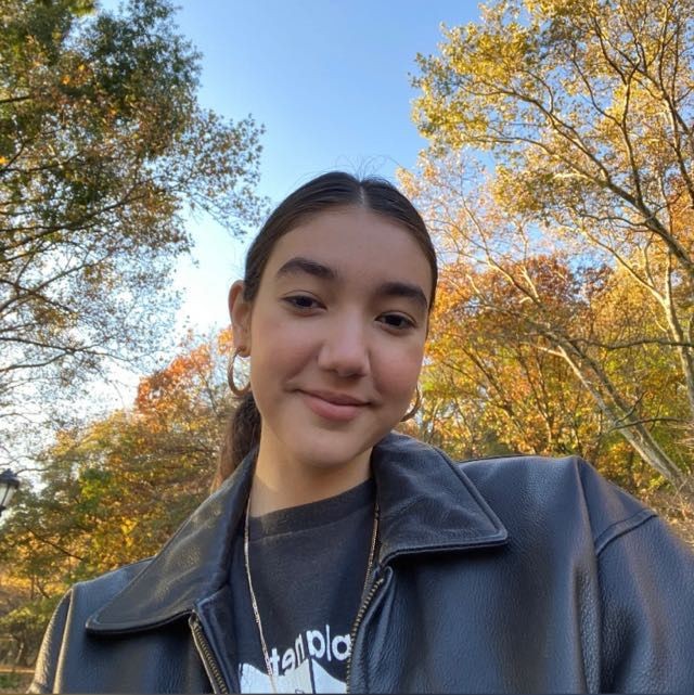 Sofia has dark brown hair, which she has on a ponytail in this picture. She is wearing a dark shirt and a black leather jacket, and golden hoop earrings. The background is the sky and some trees. 
