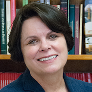 Regina Cortina smiles at camera in front of stacked bookshelf wearing a dark blazer