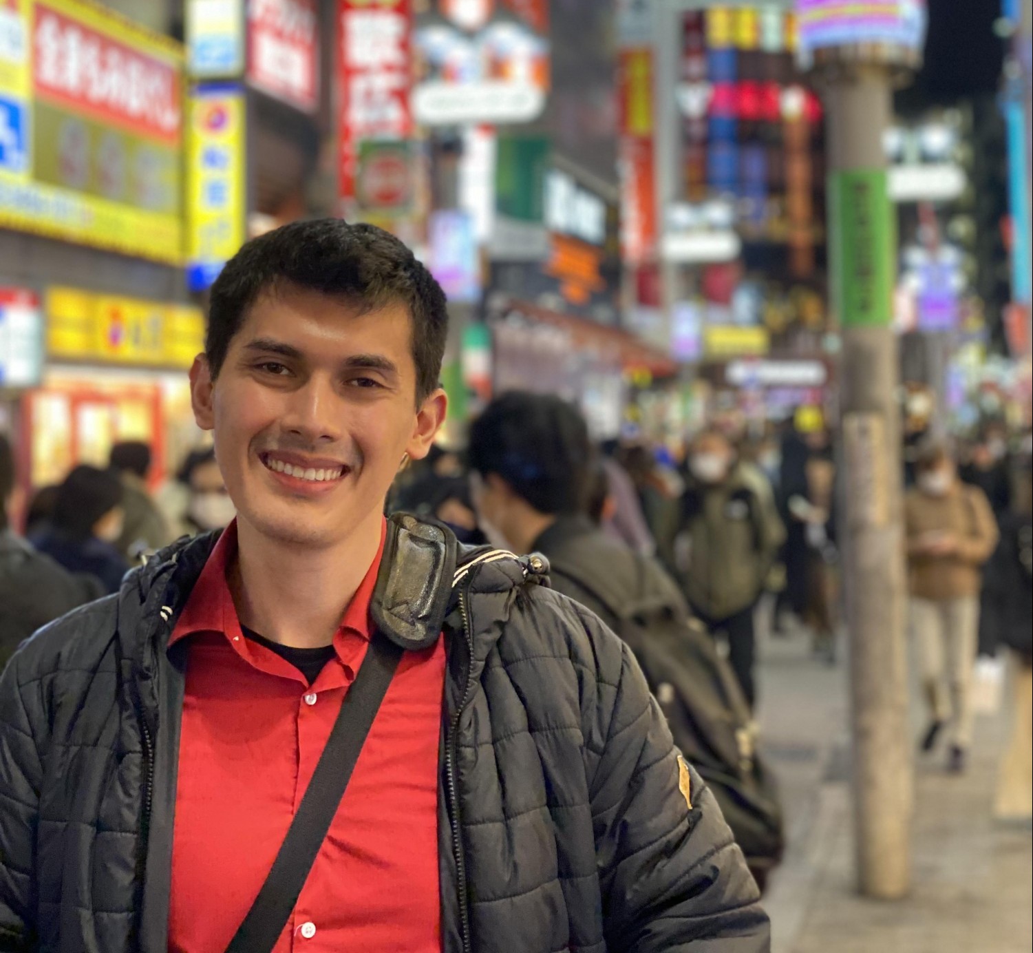 Ricardo is standing and smiling in front of a blurry background of a busy street with colorful billboards and what looks like Mandarim ideograms. He is wearing a red button-down shirt and a black thermal jacket. The black strap of a bag extends diagonally from his left shoulder to his right side.