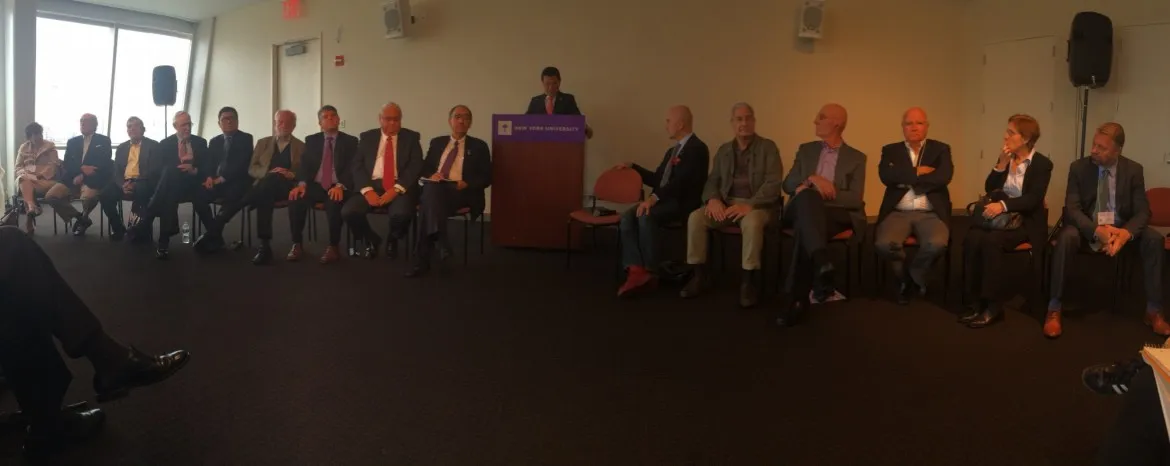 Individuals of a panel are seated horizontally across a stage while one speakers stands at the podium 