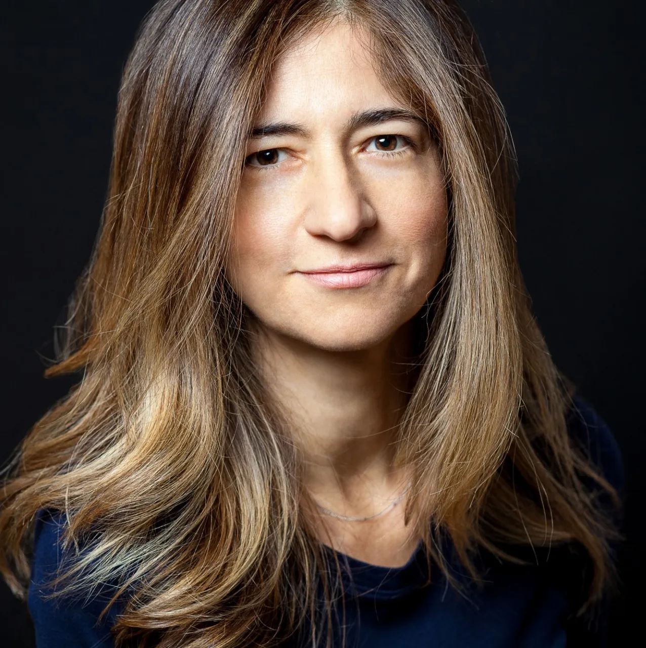 A dark-haired woman wearing a blue sweater stands in front of a dark gray background, looking directly into the camera and subtly smiling. 