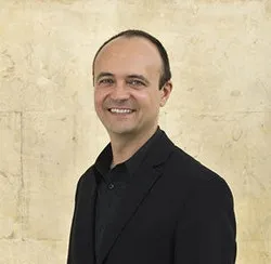 Jorge Otero-Pailos is standing in front of a wall. He wears a black suit. 