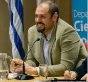 Juan Pablo Luna is smiling while sitting at a conference table. He is wearing a beige blazer and light blue shirt. On the table there is a pitcher of water, a glass filled with water, and a table microphone.