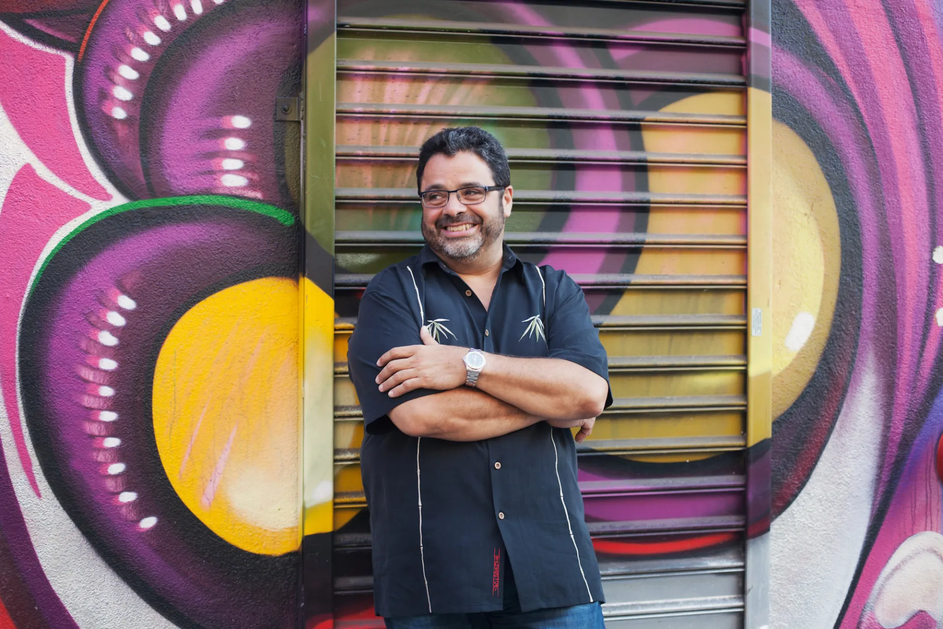 Arturo O'Farril stands in front of a colorful graffited wall. He wears a blue button up shirt. 